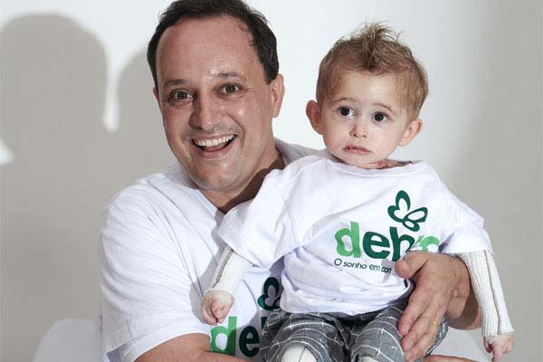 Theo, bebê com epidermólise bolhosa (EB), com curativos nos dois braços e nos joelhos, sorrindo no colo do seu tio Leandro, presidente da DEBRA
