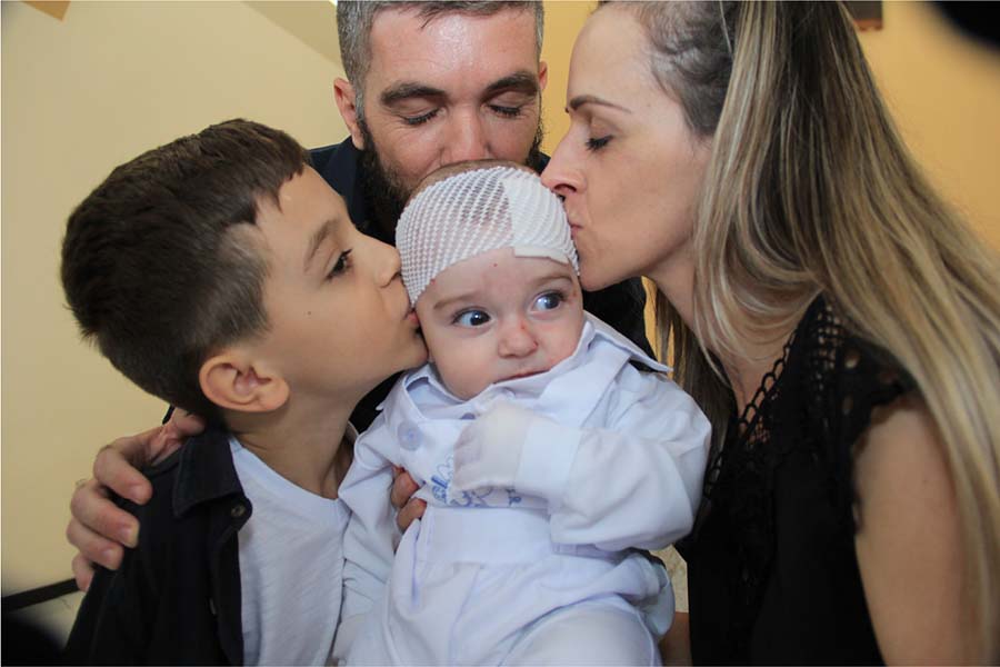 Lucas, bebê que nasceu com epidermólise bolhosa (EB), com o irmão mais velho e a mãe em cada lado dele e o pai atrás. Todos estão beijando o Lucas